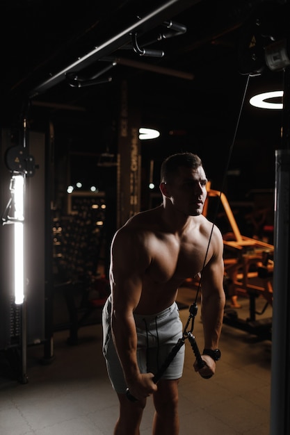 Фото Модель-бодибилдер парень с голым торсом тренируется в тренажерном зале. мышцы напряжены. темный фон.