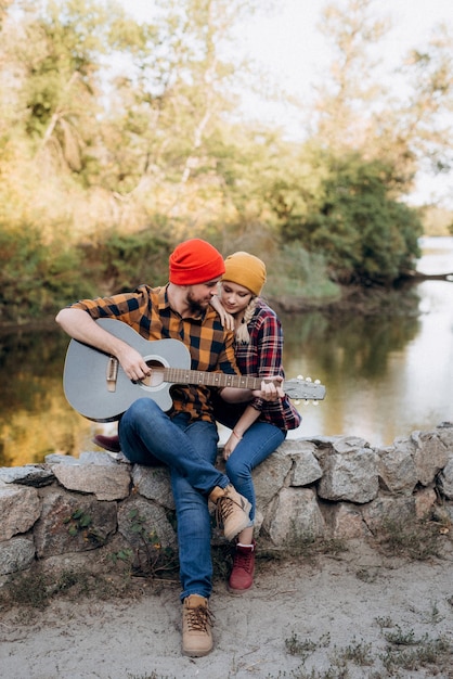 A guy in a bright hat plays the guitar with a girl against a background of granite rocks and a river