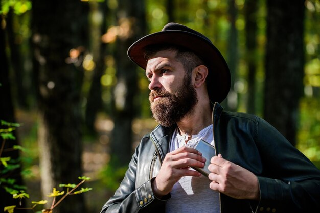 Guy brutal bearded cowboy drink alcohol metal flask. Hipster with beard drink alcohol nature background defocused. Hiking camping. Wanderer concept. Alcohol drink. Nomadic man carry alcohol with him.