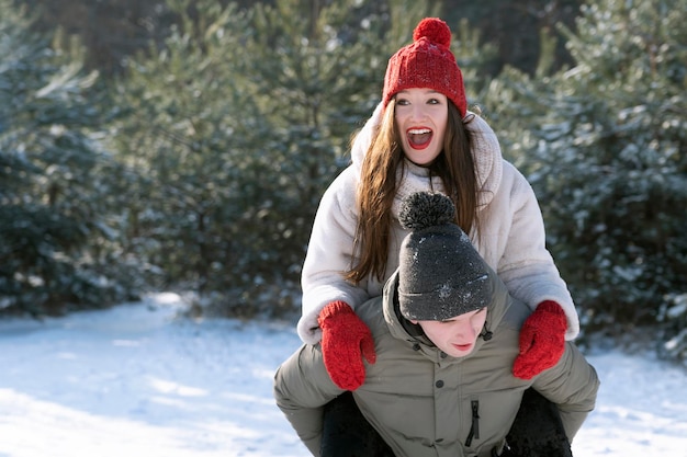 Guy carries his girlfriend on his back on winter forest background. Young couple having fun on frosty sunny day.