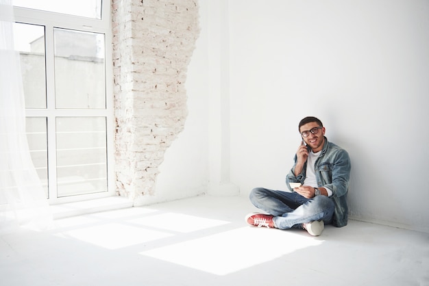 A guy in casual clothes is sitting at home in an empty apartment holding a credit card and calling on the phone. Maybe he's a new resident and has not bought any furniture yet.