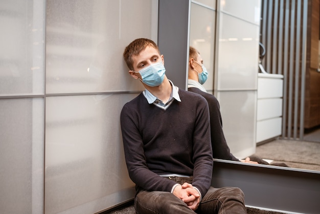 A guy of caucasian nationality sits in a corner wearing a protective mask sad at the mirror