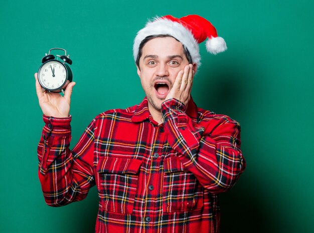 Guy in Christmas hat and tartan shirt with alarm clock on green