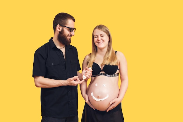 The guy draws with cream emoji on the belly of his pregnant wife on a yellow background