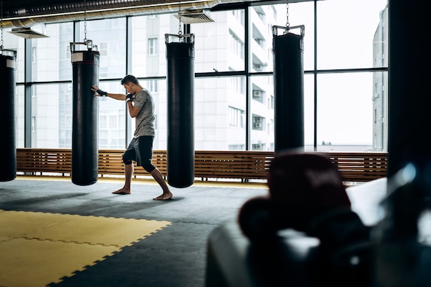 Guy dressed in the grey t-shirt and black shorts stands on guard and works out a boxing punch next to hanging punching bag against the background of panoramic windows in the gym .