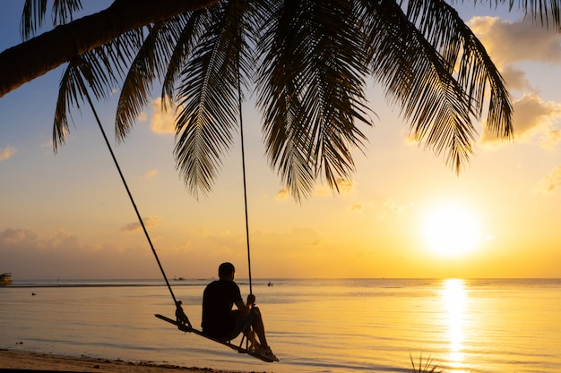 The guy enjoys the sunset riding on a swing on the ptropical beach. Silhouettes of a guy on a swing hanging on a palm tree, watching the sunset in the water.