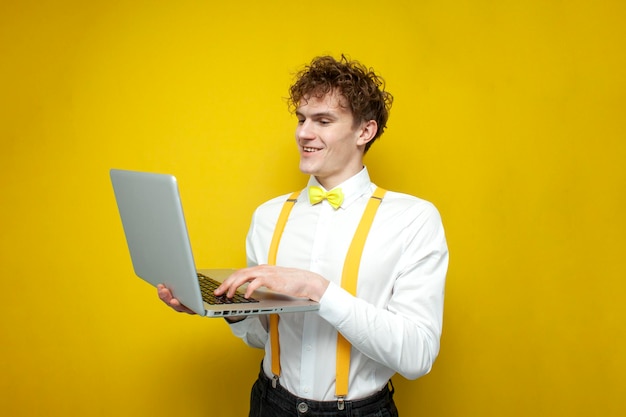Ragazzo in abito festivo usa il computer portatile su sfondo giallo isolato studente in papillon e bretelle sta scrivendo