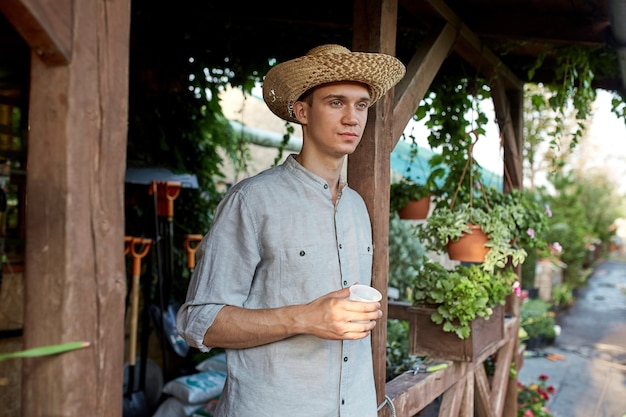 Guy gardener in a straw hat is standing with plastic glass in his hand next to a wooden veranda in the wonderful nursery-garden on a sunny day. .