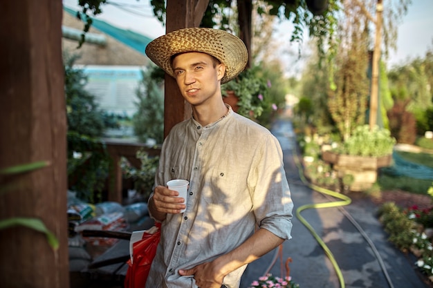 Парень-садовник в соломенной шляпе стоит с пластиковым стаканом в руке у деревянной веранды в чудесном детском саду в солнечный день. .