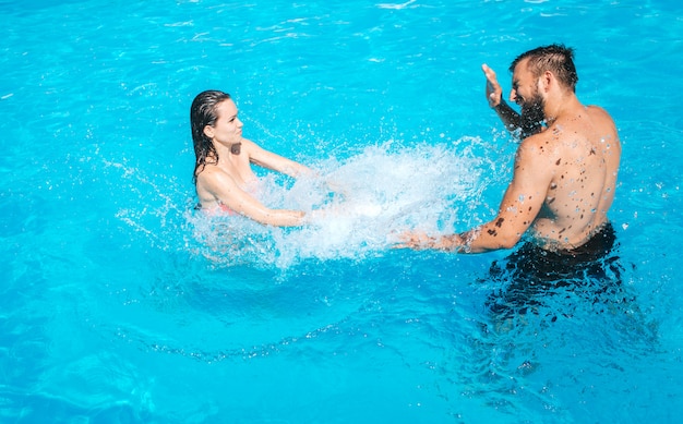 男と女はプールで遊んでいます。女の子はそこに立って、水しぶきを作ります。男は飛び散りから目を守ります。