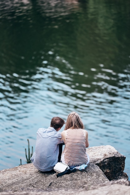 The guy and the girl are sitting on a cliff above the river and looking in front. rear view. loving couple.