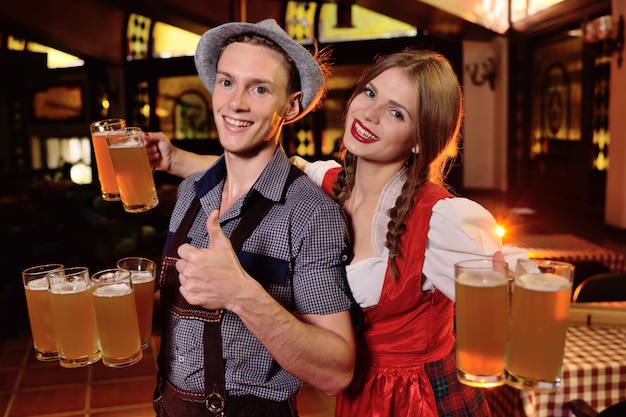 Guy and a girl in Bavarian clothes holding a lot of mugs with beer on the pub background during the celebration of Oktoberfest.