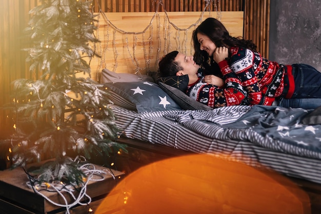 The guy and the girl on the bed gently look at each other small Christmas tree with garlands on the bedside table lens flare