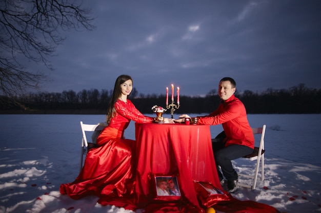 A guy and a girl in love sit in red clothes at a table with candles and hold on to their hands against a background of snow