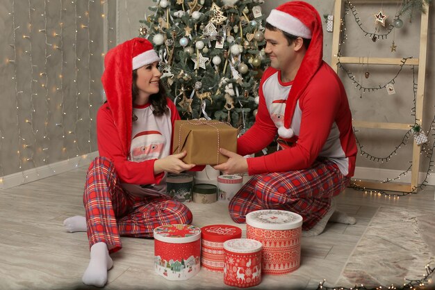 парень и девушка в пижаме и шапке Деда Мороза сидят у елки и дарят подарок