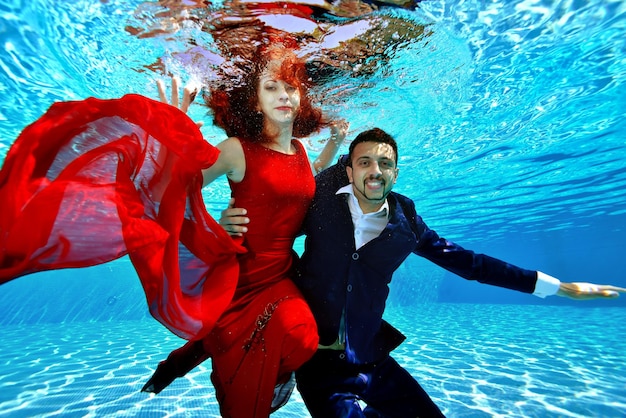 A guy and a girl in a red evening dress swim and play underwater in the pool on a Sunny day