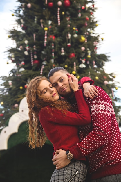 The guy and the girl in red sweaters are smiling happily at the Christmas tree.