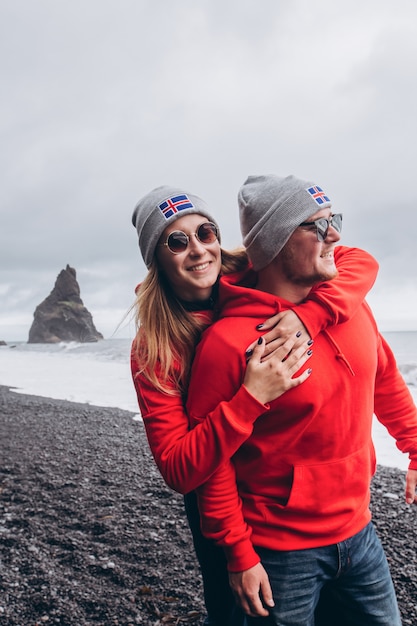 A guy and a girl in red sweaters and gray hats are hugging on a black beach, next to Vic