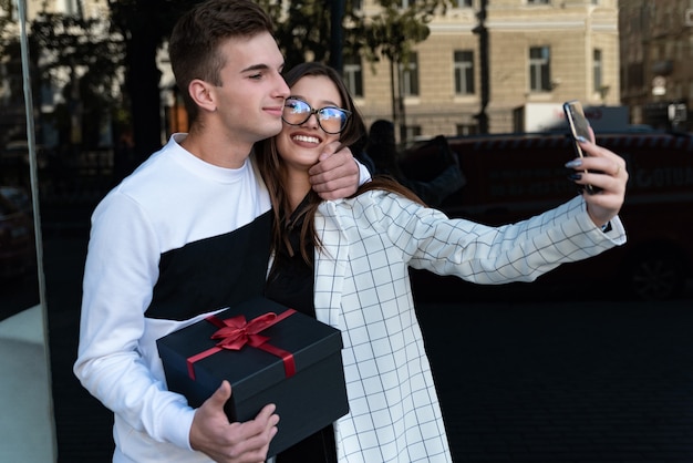 Guy gives his girlfriend a gift and hugs her. Happy couple in love with gift in their hands makes selfie. Smiling couple in love.