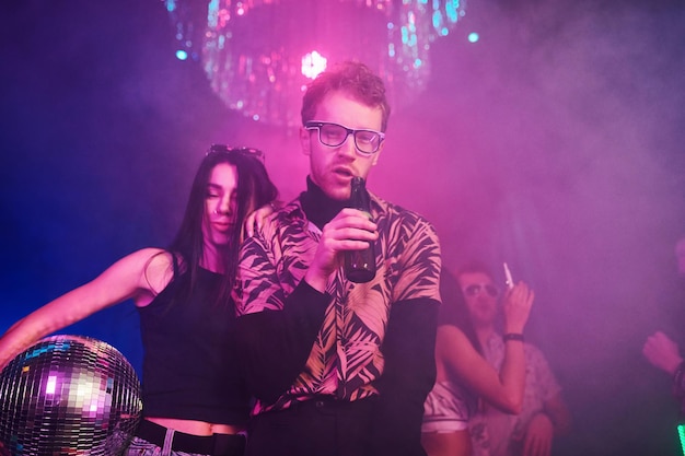 Guy in glasses drinks beer young people is having fun in night\
club with colorful laser lights