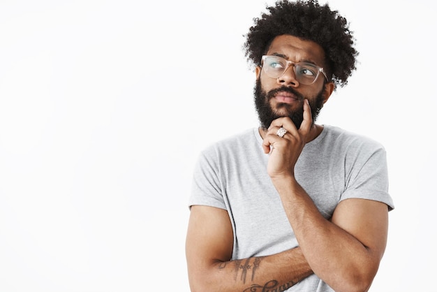 guy in glasses making plan in mind thinking up grocery list holding hand on beard looking left thoughtful, frowning as considering what choice right creating new idea on gray wall.