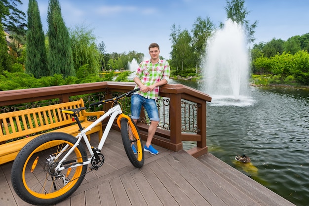 Guy in green and red plaid shirt leaning on railing standing on the wooden deck after biking in a park with fountain in a background