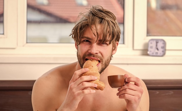 Guy having coffee and holds croissant while lay bed in bedroom\
or hotel room bearded man in bed