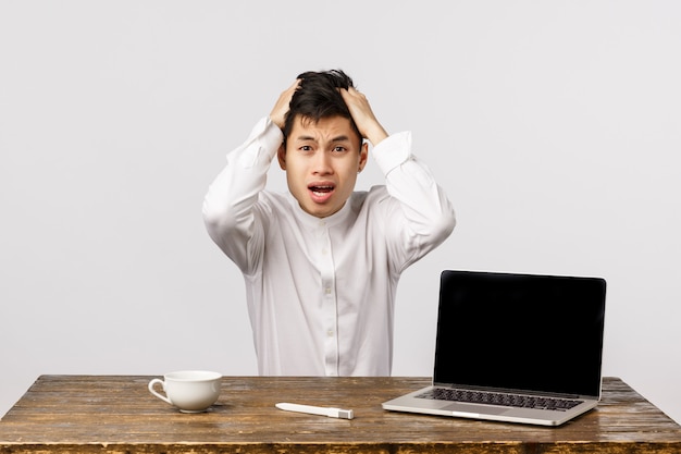 Guy having trouble, dont know what do, sitting indecisive and anxious. Shocked and distressed asian businessman panic, grab head troubled and screaming worried as react on promo laptop screen