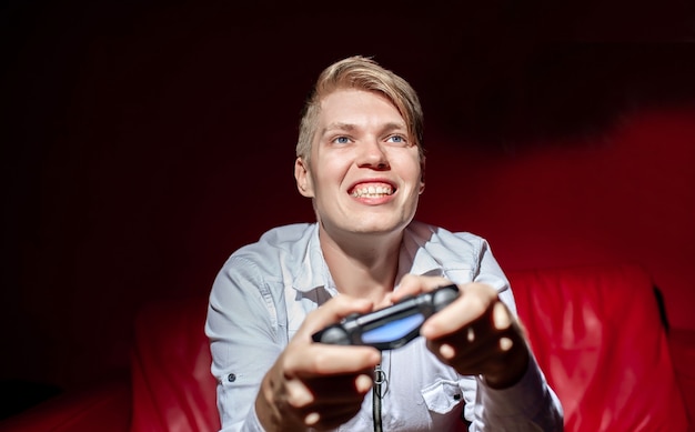 The guy holds a joystick in his hands and plays a game console, sitting on a red sofa.