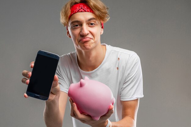 Guy holds a pink piggy bank and a smartphone with a mockup on a gray background.