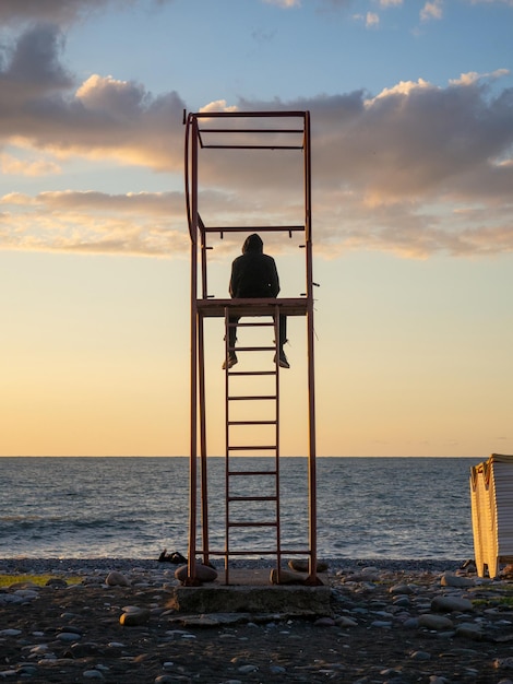 Foto tipo con il cappuccio su una torre di soccorso silhouette di un giovane uomo solitario silhouetta di una persona che guarda il mare al tramonto gambe pendenti concetto di solitudine