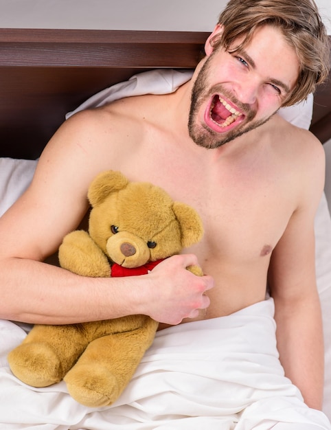 Guy hug teddy bear soft plush toy. Man unshaven bearded face relax with favorite teddy bear. Sweet dreams concept. Man handsome guy relaxing bed hug teddy bear toy. Sweet memories from childhood.