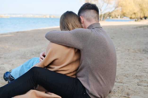 The guy hugs the girl by the shoulders The couple sits with their backs to the camera Date on the beach in the cold season