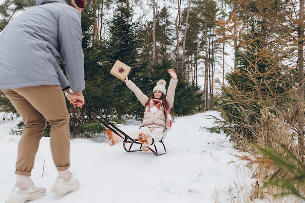 남자는 겨울 침엽수 숲에서 선물을 들고 여자 친구에게 썰매를 타며 즐거운 시간을 보내고 있습니다.