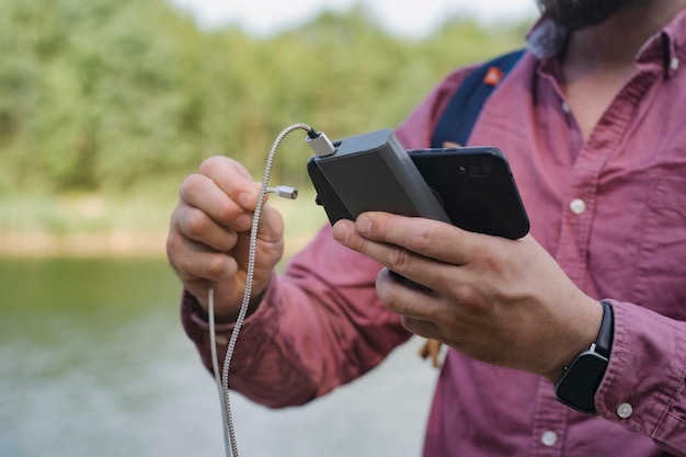 男は手にスマートフォンを持ったポータブル充電器を持っている 緑と湖のある自然の背景に男