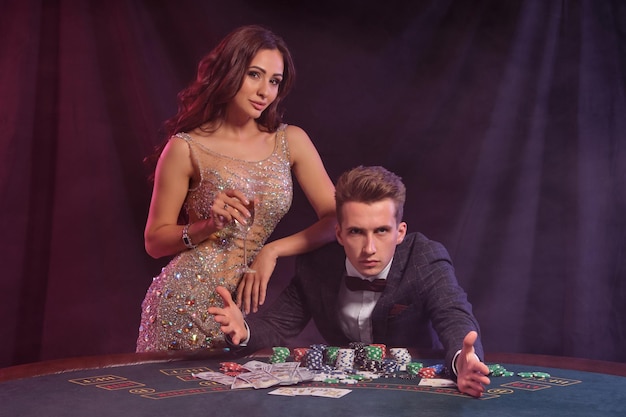 Guy is playing poker at casino at table with stacks of chips and cards on it He celebrating win with female Black smoke background Closeup