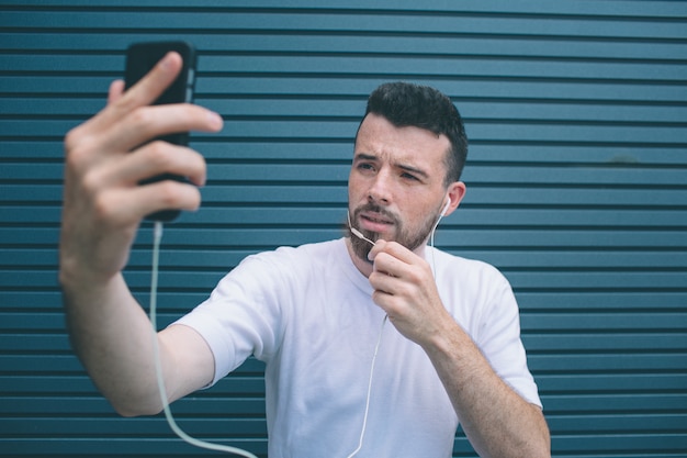 Guy is posing on camera. He is taking selfie and holding one hand on chin. Also man is listening to music. Isolated on striped 