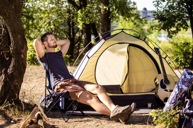 A guy is resting near a tent on a folding chair in the forest.