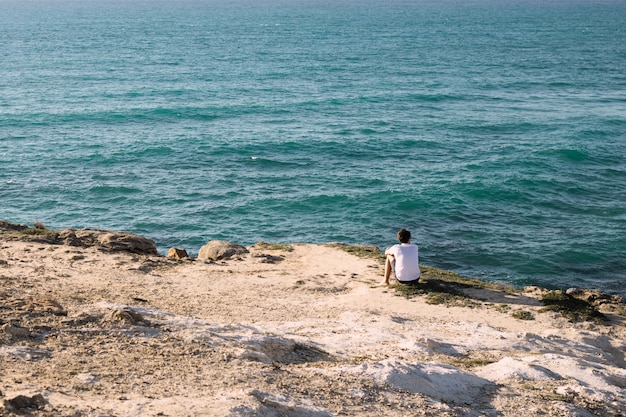 The guy is sitting on the beach by the cliff