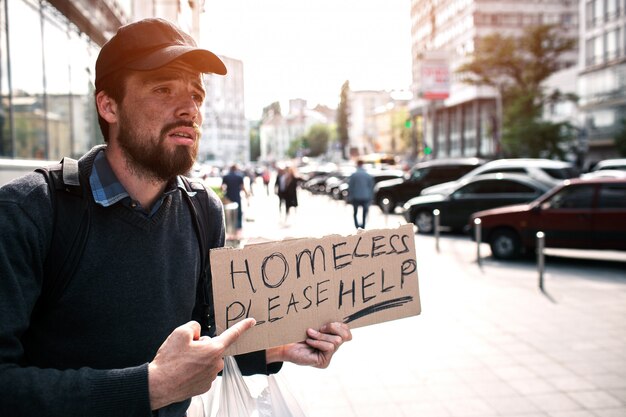 Guy is standing on the street and pointing on piece of cardboard which says homeless please help