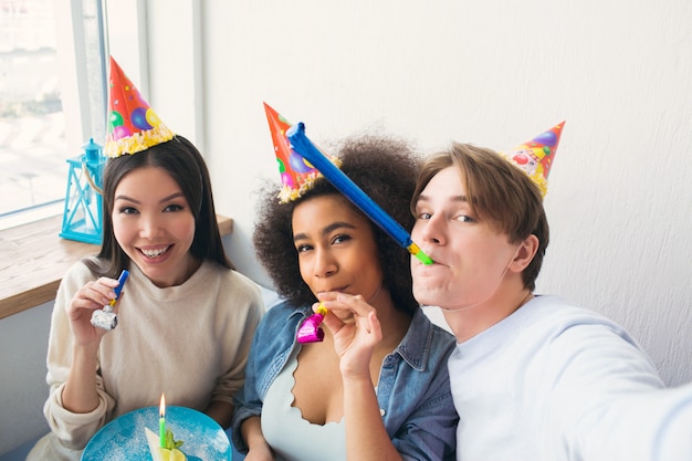 ガイは2人の友人とセルフィーを撮っています。彼らはアフロアメリカンガールズの誕生日を祝っています。人々は誕生日の帽子をかぶっています。また、女の子はwhiを持っています。