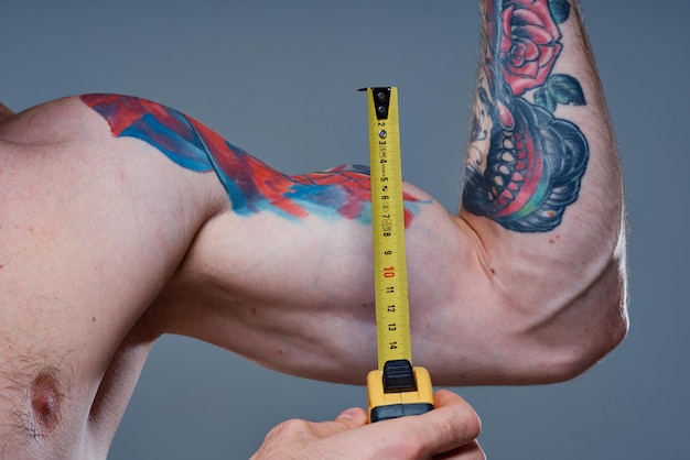 Guy measures arm muscles with centimeters on a gray background and a multicolored tattoo bodybuilder fitness