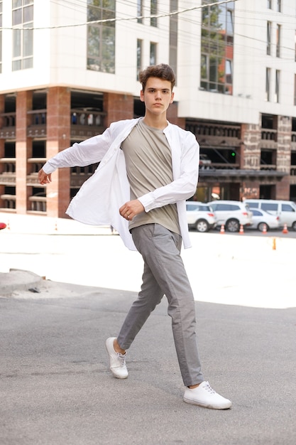 Guy model with a stylish haircut posing outdoors in a white shirt and gray trousers. trendy hairstyle rests near a modern business center. Attractive guy in the street on a summer day.