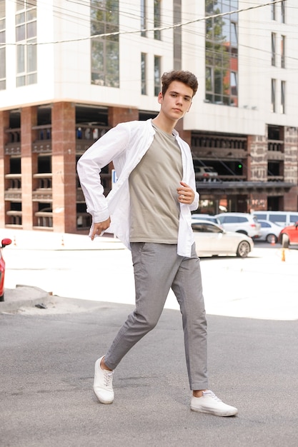 Guy model with a stylish haircut posing outdoors in a white shirt and gray trousers. trendy hairstyle rests near a modern business center. Attractive guy in the street on a summer day.