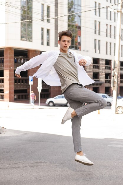 Модель парня со стильной стрижкой позирует на открытом воздухе в белой рубашке и серых брюках. модная прическа держится возле современного бизнес-центра. Молодой человек прыгает с модной одеждой против