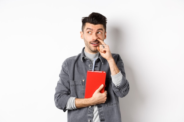 Guy picking nose and look aside at logo, holding red book or planner in hand, standing against white background.