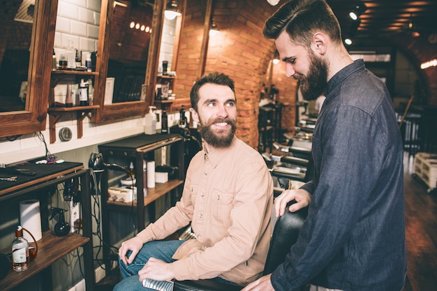 Guy in pink shirt is sitting in a chair and looking at the guy in blue shirt. He is happy to see him. Another guy is looking down to the floor.