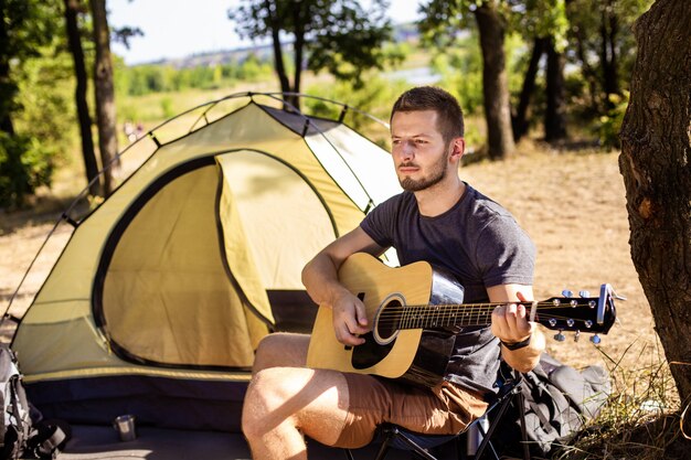 The guy plays guitar music near the tent on a folding chair in the woods