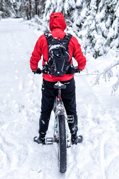 赤いジャケットを着た男が雪に覆われた森の中で自転車に背を向けて立っています