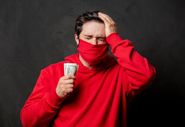 赤いスウェットシャツとフェイスマスクの男は暗い壁にお金を手に持っています